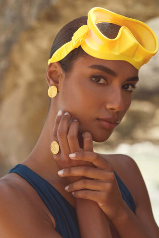 A close-up lifestyle shot of a model wearing a bright yellow swim goggles headband, gold coin earrings, and a navy blue top, set against a blurred natural rocky background.