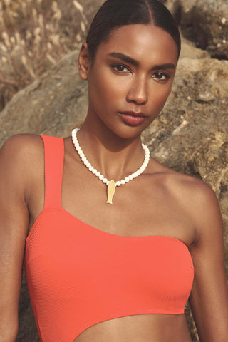 A woman wearing a coral orange one-shoulder bikini top and a white beaded necklace with a gold tooth pendant, posed outdoors against a rocky background.