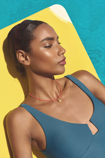 A woman in a teal swimsuit reclines on a yellow lounge chair by a swimming pool, wearing a delicate red cord necklace with a small gold pendant.
