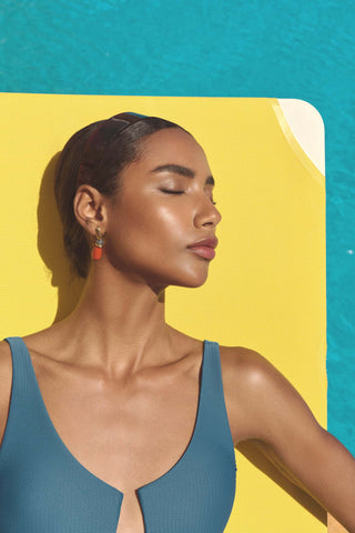 A woman in profile wearing a teal ribbed one-piece swimsuit, set against a bright yellow lounge chair and turquoise water background.