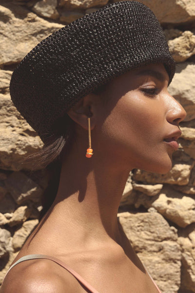 A woman in profile wearing a black woven hat and a long gold dangle earring with a small orange bead, set against a rustic stone wall.